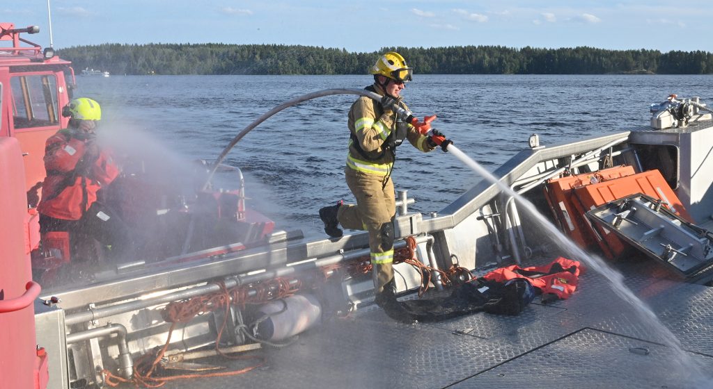 Pelastaja sammutusruiskun kanssa astumassa veneen kannelle.